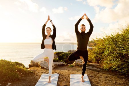 Photo for Man and woman in harmony, performing tree yoga poses on mats by the seaside, basking in the warm, glowing light of the setting sun - Royalty Free Image