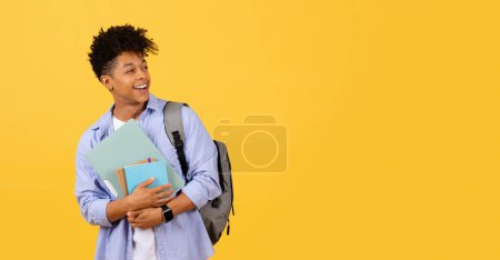 Téléchargez les photos : Jeune homme noir radieux en tenue décontractée, tenant des livres et portant un sac à dos, poutres optimistes sur fond jaune, panorama avec espace libre - en image libre de droit