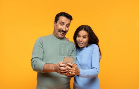 Photo for Happy shocked senior european wife and husband watch video on phone, isolated on orange studio background. Relationships, sale app, ad and offer, fun and video call surprise - Royalty Free Image