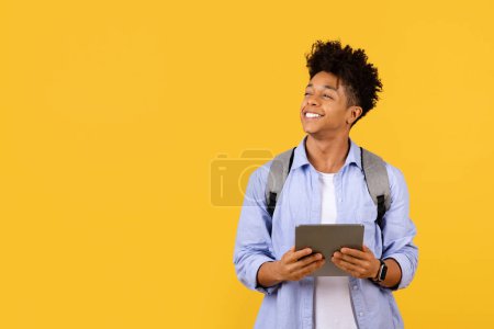 Photo for Hopeful black male student gazing upwards, holding tablet with subtle smile, suggesting aspiration and foresight on sunny yellow background, free space - Royalty Free Image