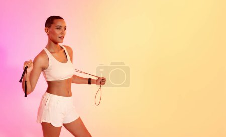 Foto de Una mujer atlética con un corte de zumbido y una expresión enfocada sostiene una cuerda de salto, lista para un entrenamiento, contra un fondo rosa y amarillo degradado. Deporte, fitness y pérdida de peso - Imagen libre de derechos