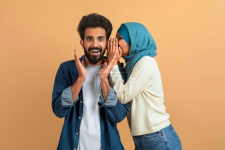 Photo for Gossips Concept. Muslim Woman In Hijab Whispering Secret To Her Surprised Boyfriends Ear, Excited Arab Man Opening Mouth In Amazement While Standing Together Over Beige Studio Background, Free Space - Royalty Free Image