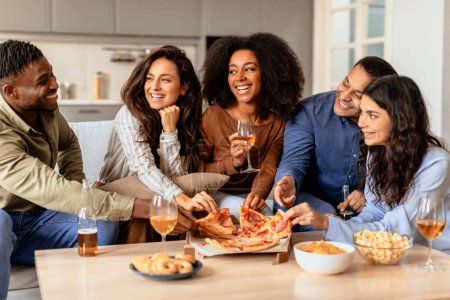 Foto de Fiesta de estudiantes. Diversos amigos se reúnen para cenar en casa con cerveza y pizza, entablando conversaciones sobre aperitivos y bebidas, compartiendo risas y alegría en una cómoda cocina interior - Imagen libre de derechos