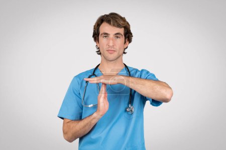 Attentive male nurse in blue scrubs with stethoscope around his neck making timeout hand gesture, indicating need for break or attention