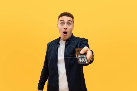 Photo for Portrait of amazed handsome guy watching TV show or film, holding remote control, switching channels, selective focus on hand. Young emotional man spending weekend free time, yellow studio background - Royalty Free Image