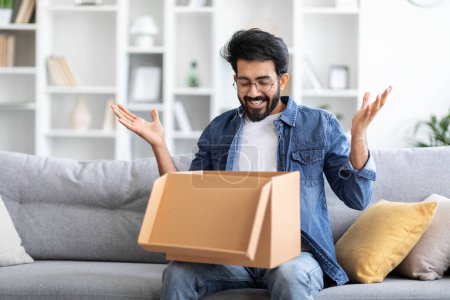 Photo for Delighted indian man expressing surprise and joy when opening cardboard package at home, happy excited eastern male unboxing parcel while sitting on couch in cozy living room, copy space - Royalty Free Image