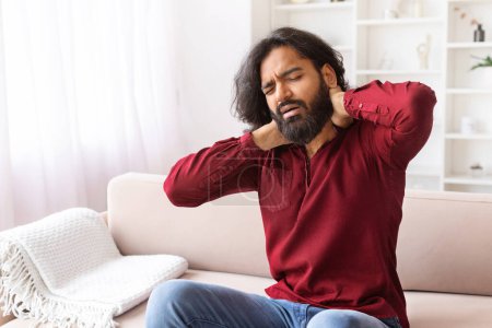 Foto de Hombre indio que sufre de dolor de cuello Masajeando músculos que sufren Sentarse en el sofá en la sala de estar en casa. Problema de Salud Neurológica, Artritis y Osteoartritis Concepto, Copiar Espacio - Imagen libre de derechos