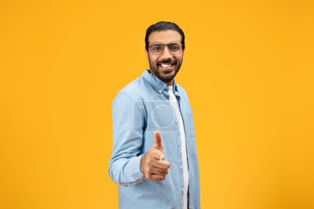 Photo for Friendly man in a denim shirt giving a thumbs up and smiling towards the camera, showcasing a sign of approval and positivity against a bright yellow background. Motivation sign - Royalty Free Image