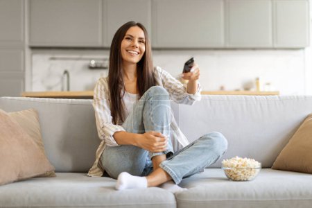 Photo for Delighted young female with bowl of popcorn at home comfortably watching her favorite tv shows, smiling woman holding remote in hand, relaxing on plush couch in living room, copy space - Royalty Free Image