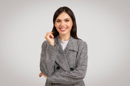 Photo for Charming young businesswoman with beaming smile, posing confidently in modern houndstooth blazer, against clean, grey backdrop - Royalty Free Image