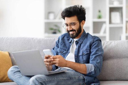 Hombre indio absorto en gafas multitarea con computadora portátil y teléfono inteligente en casa, joven freelancer oriental mensajería en el teléfono móvil y el uso de la computadora mientras está sentado en el sofá en casa