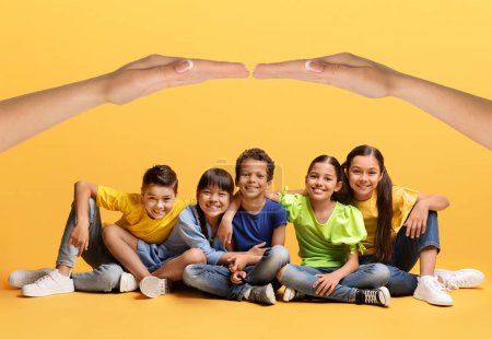 Téléchargez les photos : Heureux enfants multiethniques mignons garçons et filles en tenue décontractée assis sur le sol sur fond jaune sous le toit composé de deux mains. Sécurité enfance, protection, assurance, soins - en image libre de droit
