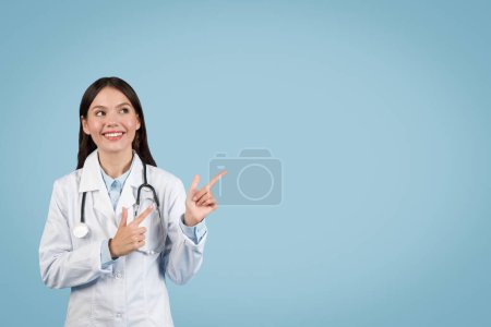 Fröhliche Ärztin zeigt nach oben in den freien Raum, schaut auf etwas Interessantes, mit Stethoskop um den Hals, auf frischem blauen Hintergrund