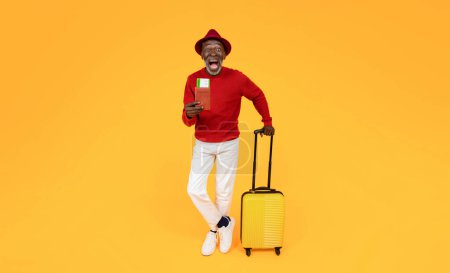 Foto de Feliz emocionado hombre afroamericano senior turista en sombrero con maleta amarilla y la boca abierta mostrar pasaporte y boletos de avión, listo para viajar, aislado en fondo de estudio naranja. Viaje, aventura - Imagen libre de derechos
