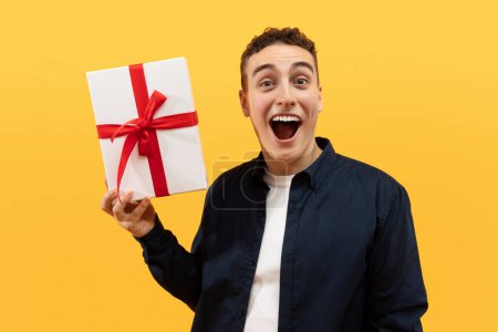 Photo for Excited cool funny handsome blonde young guy wearing casual clothing holding white gift box with red bow and grimacing, celebrating birthday on yellow studio background - Royalty Free Image