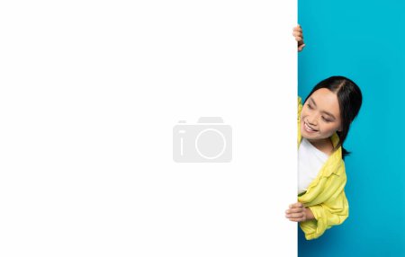 Photo for An Asian millennial woman with a joyful expression is playfully peeking from behind a blank white vertical banner, providing ample space for text on a turquoise background - Royalty Free Image