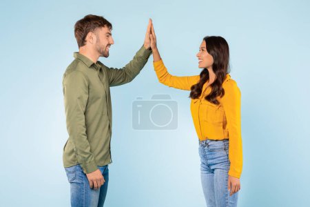 Téléchargez les photos : Souriant jeune homme et femme en tenue colorée joyeusement haut-cinq l'un l'autre, symbolisant le travail d'équipe et le succès sur fond bleu vif, vue de côté - en image libre de droit