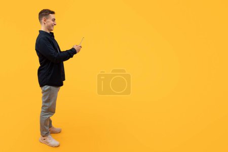 Photo for Mobile application for communication. Stylish cheerful young caucasian guy wearing casual clothing using smartphone isolated on yellow studio background, texting friends, scrolling, copy space - Royalty Free Image