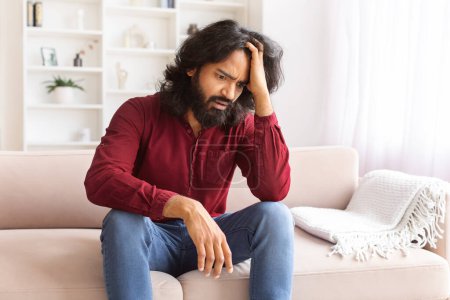 Photo for Distressed millennial bearded indian man sitting on couch alone, touching head, feeling down, experiencing breakup divorce, lost job, going through financial or personal crisis, copy space - Royalty Free Image