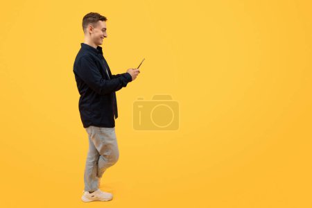 Téléchargez les photos : Application mobile. Jeune homme élégant en vêtements décontractés utilisant un téléphone portable sur fond de studio jaune, websurf, shopping ou banque en ligne, textos amis, pleine longueur, espace de copie - en image libre de droit