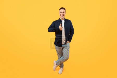 Photo for Positive stylish handsome young caucasian guy wearing casual clothing showing thumb up and smiling at camera, recommending nice offer, yellow studio background, full length, copy space - Royalty Free Image