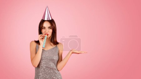 Téléchargez les photos : Femme ludique dans une robe pétillante et chapeau de fête soufflant une corne de fête, avec une main tendue comme pour montrer quelque chose, sur un fond rose festive. Événement de fête - en image libre de droit