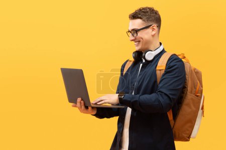 Téléchargez les photos : Jeune étudiant cool avec sac à dos et écouteurs sans fil portant des lunettes à l'aide d'un ordinateur portable sur fond de studio jaune, espace de copie. Éducation en ligne, e-learning - en image libre de droit