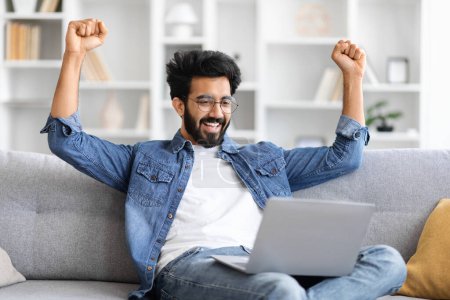 Téléchargez les photos : Joyeux homme indien en chemise en denim levant les poings dans la victoire tout en regardant l'écran d'ordinateur portable, heureux gars de l'Est assis sur le canapé dans le salon confortable, célébrant le succès et la réussite personnelle - en image libre de droit