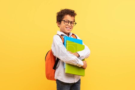 Foto de Niño adolescente negro inteligente con gafas que sostienen carpetas de colores y llevan mochila, listo para la escuela, se levanta contra el fondo amarillo brillante - Imagen libre de derechos