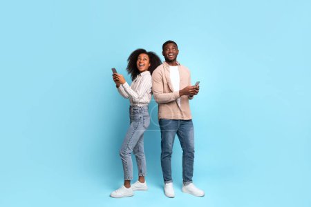 Fröhliches junges afrikanisch-amerikanisches Paar, Rücken an Rücken stehend, mit entzücktem Lächeln in ihre Smartphones vertieft, vor leuchtend blauem Hintergrund