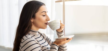 A serene young woman with long dark hair, wearing a striped turtleneck, enjoys a moment of tranquility while sipping coffee from a white cup, closing her eyes against a bright window