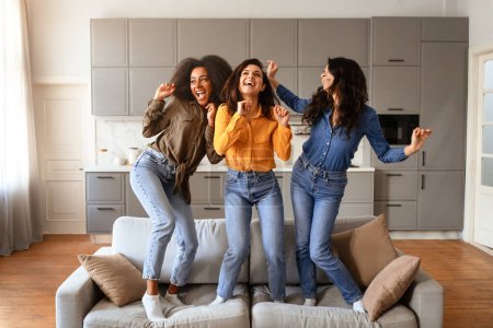 Foto de Tres señoritas multirraciales riendo y bailando juntas de pie en el sofá, durante una animada fiesta de fin de semana en el salón interior, haciendo movimientos divertidos y divirtiéndose - Imagen libre de derechos