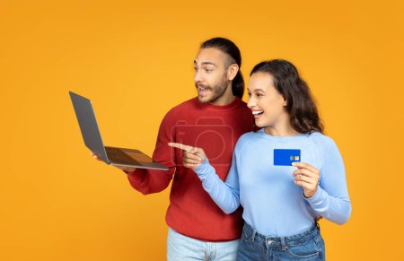 Photo for Excited young couple with bank credit card and laptop computer in their hands posing on orange studio background. Spouses shopping or banking online, got cashback - Royalty Free Image