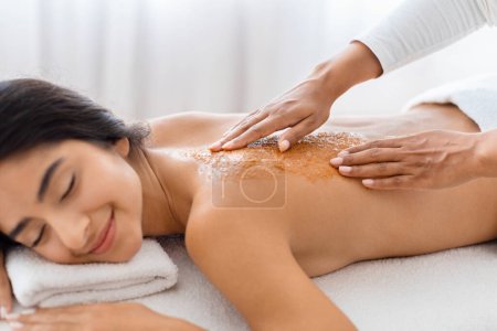Photo for Serene relaxed young indian woman deeply relaxing as a spa professional applies an exfoliating treatment to her back, promoting wellness and skin health, closeup - Royalty Free Image