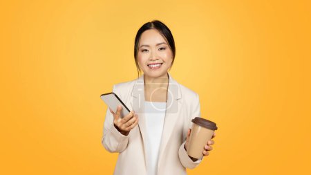 Photo for Glad millennial chinese lady in suit with cup of takeaway coffee, chatting on smartphone, isolated on yellow studio background, panorama. Work app, ad and offer, lifestyle - Royalty Free Image