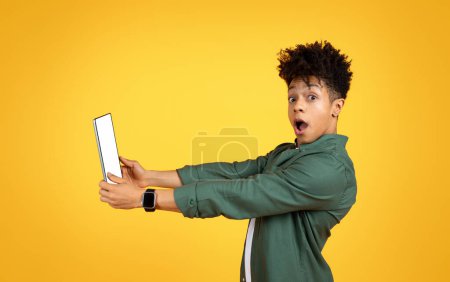 Photo for Exciting online offer. Amazed emotional young black guy holding digital tablet with white blank screen and looking at camera, checking nice mobile app, yellow background, blank space - Royalty Free Image