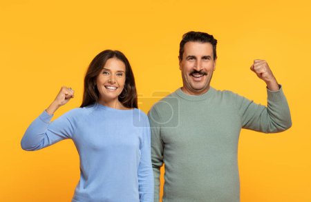 Téléchargez les photos : Heureux séniors gagnants du couple européen lèvent les poings dans un geste triomphant, souriant avec enthousiasme et confiance, sur fond orange. Succès et confiance, victoire à célébrer - en image libre de droit