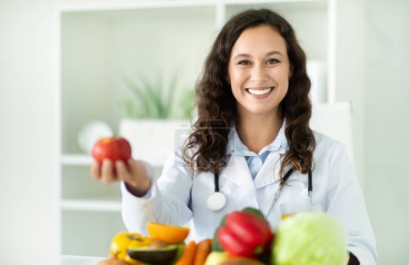 Téléchargez les photos : Jeune diététiste européenne tenant une pomme, assise au bureau avec des fruits et légumes, souriant à la caméra. Lady nutritionniste recommandant clinique de perte de poids, espace de copie - en image libre de droit