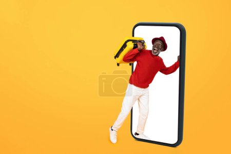 Téléchargez les photos : Joyeux homme âgé noir en pull rouge et chapeau rouge sortant d'un smartphone géant, portant une valise jaune, incarnant des thèmes de voyage et de technologie sur un fond jaune vif - en image libre de droit