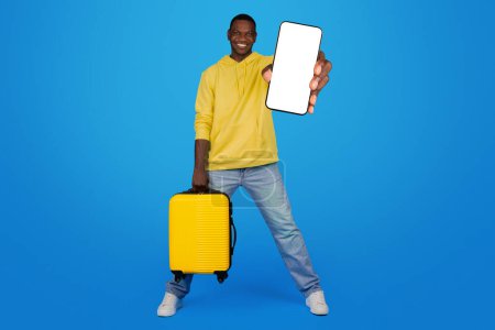 Photo for An exuberant Black man strides forward with a yellow suitcase, holding up a smartphone with a mockup screen, against a lively blue backdrop, exuding travel readiness and joy - Royalty Free Image
