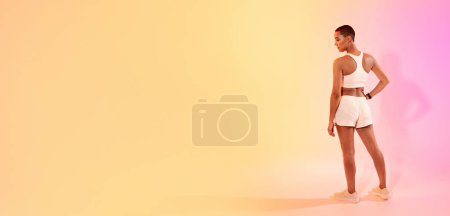 Téléchargez les photos : Une femme contemplative avec une tête rasée regarde vers le côté tout en se tenant debout dans un soutien-gorge de sport blanc et short, présenté sur un fond de dégradé doux, panorama. Fit, sport et soins de santé - en image libre de droit