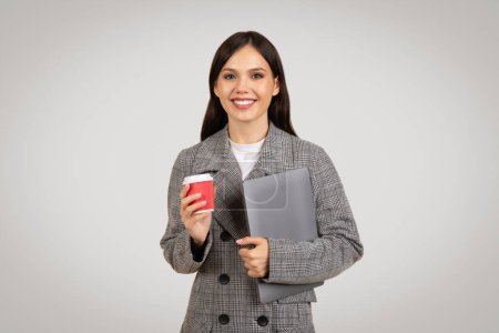 Téléchargez les photos : Femme d'affaires souriante dans un blazer à dents de poule tenant une tasse de café rouge et un ordinateur portable gris, prête à travailler, sur un simple fond gris clair - en image libre de droit