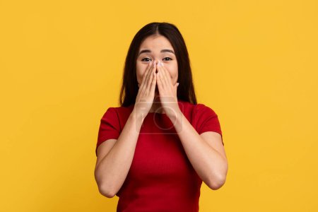 Photo for Surprised amazed emotional pretty long-haired young chinese woman wearing red t-shirt cover her mouth, touching face and looking at camera, isolated on yellow wall background - Royalty Free Image