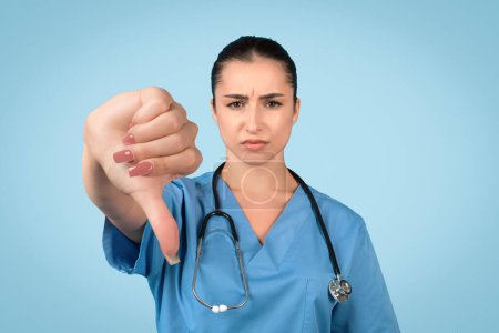 Female nurse in somber mood displays thumbs-down gesture with her hand on blue background, symbolizing disappointment in healthcare