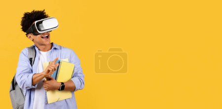 Foto de Joven estudiante masculino absorto experimentando realidad virtual con auriculares VR, libros en la mano, expresando alegría en un entorno de aprendizaje inmersivo en amarillo, panorama con espacio para copiar - Imagen libre de derechos