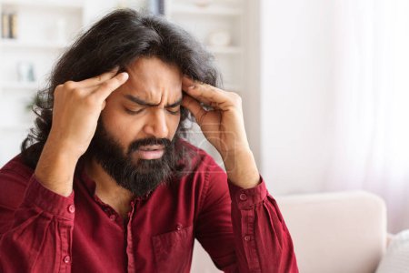 Photo for Closeup of unhappy bearded millennial indian man have strong headache, sitting on couch with closed eyes, rubbing temples, suffering from migraine, home interior, copy space - Royalty Free Image