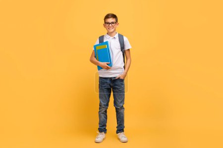 Portrait of smiling schoolboy with backpack and workbooks posing over yellow background in studio, happy teen male child in eyeglasses looking at camera, ready to school, enjoying study, full length