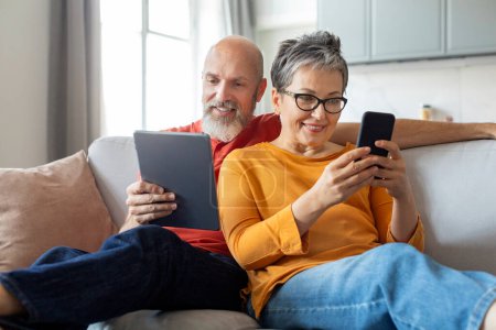 Téléchargez les photos : Heureux conjoints aînés se détendre avec des gadgets modernes sur le canapé à la maison, Souriant couple âgé reposant dans le salon intérieur, Homme utilisant la tablette numérique tandis que la femme naviguant App sur Smartphone - en image libre de droit