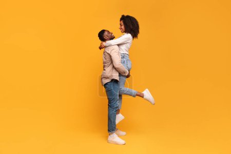 Téléchargez les photos : Couple noir heureux en vêtements décontractés s'engageant dans le dos de piggyback ludique et affectueux, avec la femme levée et souriante, sur fond jaune vif - en image libre de droit