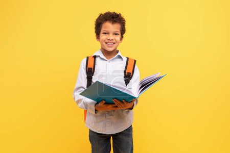 Foto de Escolar negro inteligente absorto en el libro de lectura, con una mochila naranja sonriendo a la cámara sobre un fondo amarillo vibrante - Imagen libre de derechos
